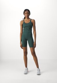 Grønn sportstanktop og tettsittende shorts laget av glatt stoff. Modell iført grå sneakers står mot en hvit bakgrunn.