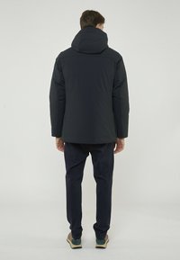 Giubbotto puffer blu navy con cappuccio, caratterizzato da una texture liscia, una vestibilità aderente e un design semplice. Indossato con pantaloni scuri e scarpe casual.