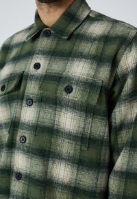 Zaļš un kr�ēmkrāsas šallveidīgs flaneles krekls ar melniem pogām, divām krūšu kabatām, punktētu apkakli un mīkstu, teksturētu audumu.