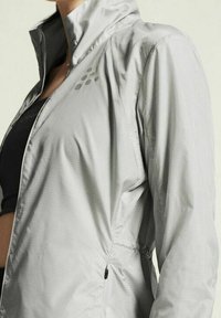 Veste coupe-vent argentée légère avec fermeture éclair, motif texturé, col haut et taille resserrée pour un ajustement réglable.