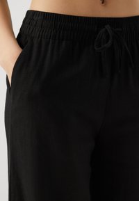 Pantalones negros de cintura elástica con cordón, que incluyen bolsillos laterales y están hechos de una tela ligera, mostrados desde la cintura hasta la mitad del muslo.