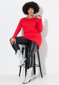 Rotes langärmliges Oberteil, schwarze glänzende Leggings und silberne Cowboystiefel. Das Model posiert auf einem schwarzen Hocker, hält Sonnenbrillen in der Hand und hat einen verspielten Gesichtsausdruck.