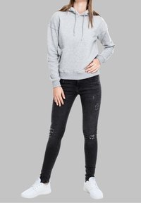 Persona vistiendo una sudadera gris claro, jeans ajustados negros desgastados y zapatillas blancas, de pie con una mano en la cadera contra un fondo gris.