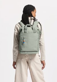 Vihreä rullattava reppu, joka on valmistettu kestävästä materiaalista. Repussa on edessä vetoketjullinen tasku, jossa on sinisiä ja oransseja yksityiskohtia, sekä pehmustetut olkahihnat.