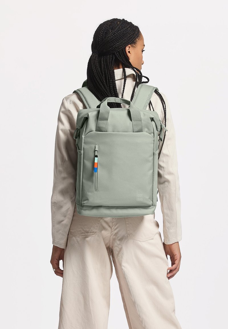 Vihreä rullattava reppu, joka on valmistettu kestävästä materiaalista. Repussa on edessä vetoketjullinen tasku, jossa on sinisiä ja oransseja yksityiskohtia, sekä pehmustetut olkahihnat.