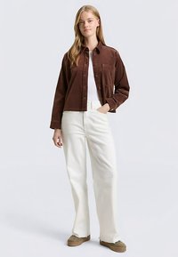 Bruine corduroy jas met knoopsluiting, een enkele borstzak, wit katoenen T-shirt en witte wijde broekspijpen. Het model draagt bruine schoenen.