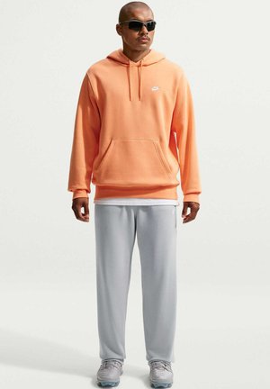 Hombre con sudadera con capucha naranja de Nike, pantalones deportivos gris claro, zapatillas grises y gafas de sol negras, posando frente a un fondo liso.