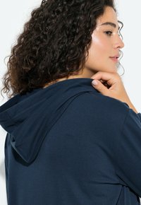 Granatowa bluza z kapturem wykonana z miękkiego materiału, wyposażona w kaptur ze sznurkiem i luźny krój. Tekstura wydaje się gładka, z minimalną liczbą widocznych szwów.