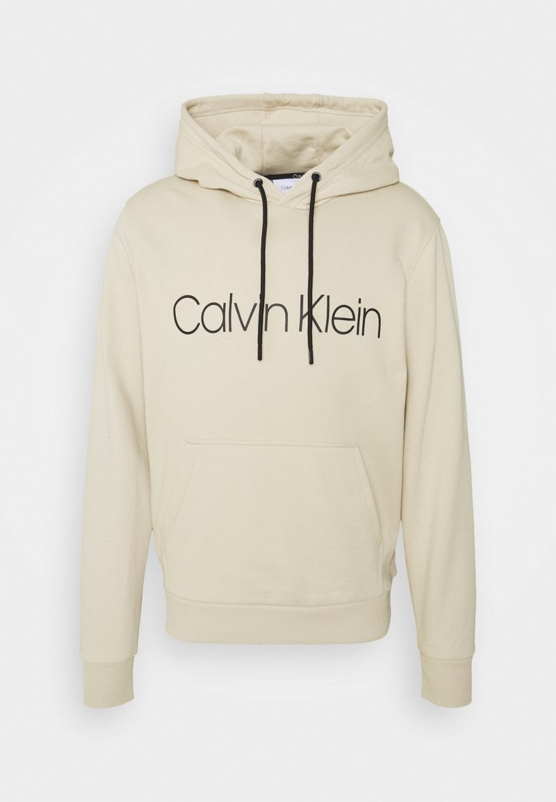Sudadera ligeramente beige con cordones negros y un gran logo de "Calvin Klein" en el frente. Cuenta con un bolsillo canguro y puños acanalados.