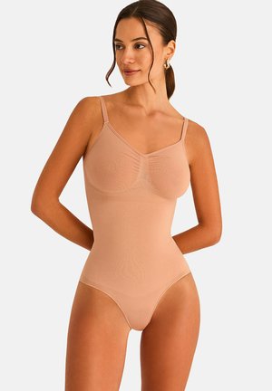 Femme aux cheveux foncés attachés en queue de cheval, portant un body nude sans couture avec des bretelles réglables, regardant vers sa gauche sur un fond uni.