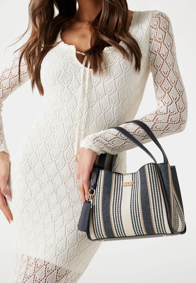 Femme portant une robe en dentelle blanche tenant un sac à main rayé noir et beige avec des poignées noires et des accents dorés.