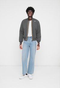 Παρκά jacket σε γκρι χρώμα με κίτρινες φερμουάρ, συνδυασμένο με ανοιχτό μπλε παντελόνι σε φαρδύ σχέδιο και λευκά αθλητικά παπούτσια, σε απλό φόντο.