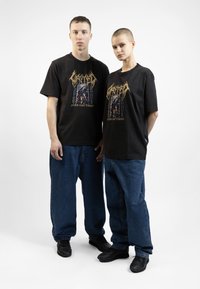 Juoda grafika t-shirts su dideliu dizaino elementu, derinamas su laisvais mėlynais džinsais ir juodais sportbačiais. Paprastas, kasdieniškas stilius.