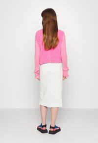 Gilet en crochet rose sur un haut à manches longues en rib (côtes), jupe midi à rayures blanches et sandales noires épaisses avec des accents colorés.
