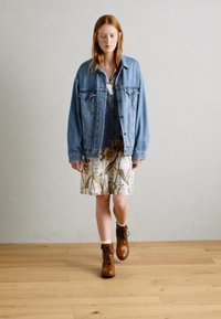 Une veste en jean, une jupe à fleurs avec un fond clair, des bottes en cuir brun à lacets et un haut superposé forment une tenue décontractée avec des textures et des couleurs variées.