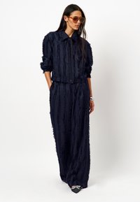 Marineblauwe twee-delige set gemaakt van gestructureerde stof met verticale strepen. De blazer heeft een kraag en een knoopsluiting; de broek is wijd uitlopend.