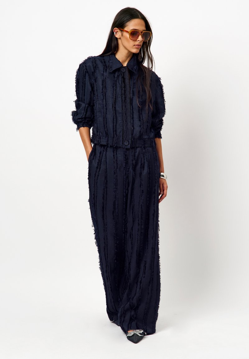 Marineblauwe twee-delige set gemaakt van gestructureerde stof met verticale strepen. De blazer heeft een kraag en een knoopsluiting; de broek is wijd uitlopend.