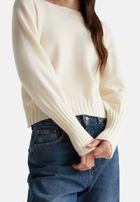 Maglione crema con polsini e orlo a coste, abbinato a jeans blu a vita alta. Struttura in maglia evidente, vestibilità casual, design minimal.