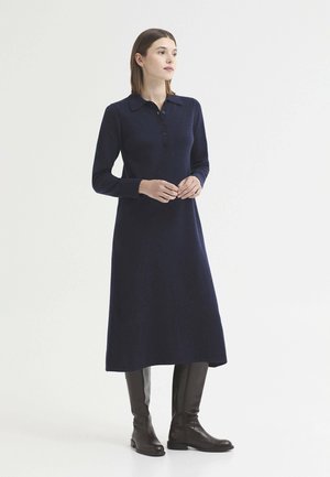 Femme debout portant une robe bleu marine à manches longues avec col et des bottes noires montantes jusqu'aux genoux sur un fond blanc uni.