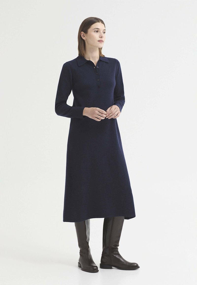 Femme debout portant une robe bleu marine à manches longues avec col et des bottes noires montantes jusqu'aux genoux sur un fond blanc uni.