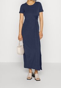 Granatowa, krótko rękawowa sukienka maxi z skręconym detalem w talii; zestawiona z beżowymi klapek na słupku i małą torebką.