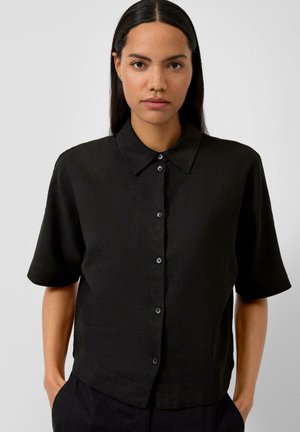 Femme aux cheveux raides et foncés porte une chemise noire à manches courtes avec des boutons, debout les mains dans les poches devant un fond uni.