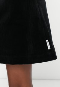 Svart kordfløyelskjørt med vertikal ribbing, tettsittende design og en liten hvit etikett ved kanten. Glatt tekstur og midtlengde.