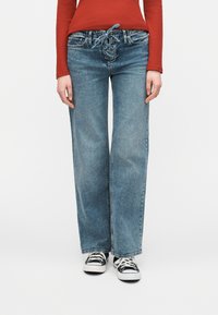 Άτομο που φοράει μπλε παντελόνι με φαρδιά μπατζάκια και κορδόνια μπροστά, και μαύρα και άσπρα αθλητικά παπούτσια, σε συνδυασμό με μακρυμάνικη κόκκινη μπλούζα.