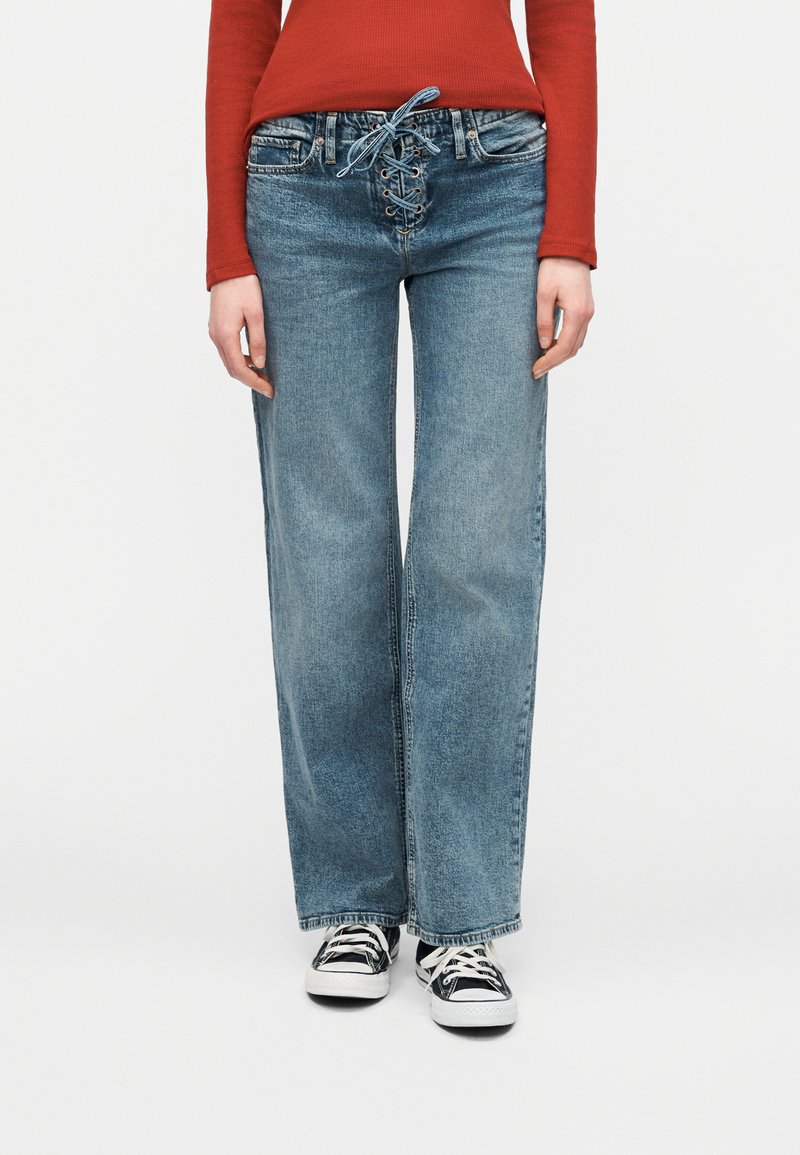 Άτομο που φοράει μπλε παντελόνι με φαρδιά μπατζάκια και κορδόνια μπροστά, και μαύρα και άσπρα αθλητικά παπούτσια, σε συνδυασμό με μακρυμάνικη κόκκινη μπλούζα.