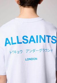 Balta medvilnės marškinėliai su „ALLSAINTS“ užrašytu ryškiu mėlynu tekstu nugaroje, papildomu tekstu japonų kalba ir „LONDON“ žemiau.