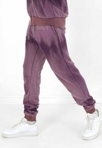Lila Trainingshose mit geripptem Bündchen und Säumen. Sie verfügt über seitliche Taschen und ein dezentes Logo am Oberschenkel. Kombiniert mit weißen Sneakers.