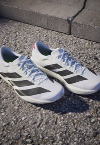 Weiße Laufschuhe mit schwarzen Streifen und hellblauen Schnürsenkeln auf rauem Asphalt nahe einem Betonbordstein.
