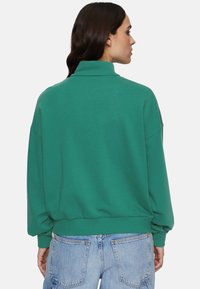 Felpa verde oversize con collo alto, spalle ampie e orlo a costine. Abbinata a shorts in denim azzurro chiaro.
