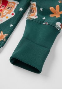 Het lange mouwen shirt heeft een groene stof met patronen van een huis van peperkoek en sterren. De manchet is geribbeld, wat zorgt voor een strakke pasvorm en een zachte textuur.