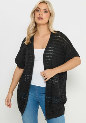 Femme portant un cardigan noir à manches courtes en maille ajourée sur un débardeur blanc et un jean bleu, se tenant devant un fond uni.