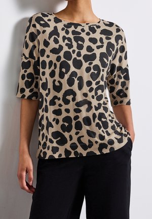 Beżowa bluzka z krótkim rękawem w nieregularny czarny wzór lamparta, noszona z czarnymi spodniami, osoba pozująca z jedną ręką w kieszeni.