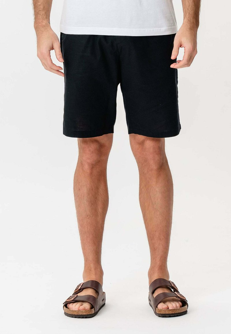 Mand iført sorte shorts, hvid skjorte og brune sandaler med to stropper, der står foran en ensfarvet lyse baggrund.