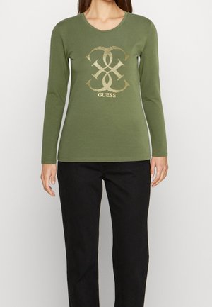 Kobieta ubrana w oliwkowo-zieloną bluzkę z długim rękawem z złotym logo GUESS oraz czarne spodnie, stojąca na jasnym, jednolitym tle.