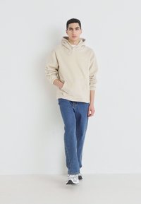 Beige hettegensere med frontlomme og justerbar snor, parret med blå rett ben jeans og hvite sneakers, på en ensfarget bakgrunn.