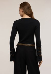 Femeie îmbrăcată într-un top negru cu mâneci lungi, având decupaje în formă de diamant pe manșetele evazate și pantaloni negri cu un săculeț maro cu ținte, întoarsă cu spatele.