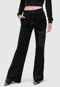 Schwarze Velour-Hosen mit weitem Bein, ausgestattet mit einem Kordelzugbund und seitlichen Taschen. Kristallakzente formen das Wort "Juicy" am linken Bein.