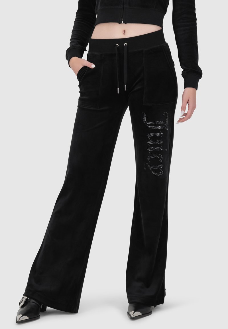 Schwarze Velour-Hosen mit weitem Bein, ausgestattet mit einem Kordelzugbund und seitlichen Taschen. Kristallakzente formen das Wort "Juicy" am linken Bein.