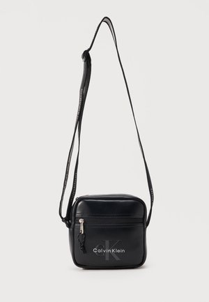 Small square black crossbody bag with front zipper pocket, adjustable strap, and Calvin Klein logo on front.