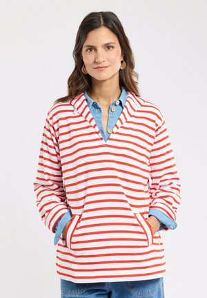 Femme aux cheveux foncés portant des boucles d'oreilles en or, un sweat à capuche blanc à rayures rouges sur une chemise bleue boutonnée, les mains dans les poches avant.