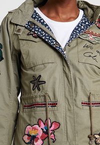 Veste verte avec broderies florales et étoile de mer, doublure intérieure bleue à motifs et taille à cordon, portée sur une chemise blanche.