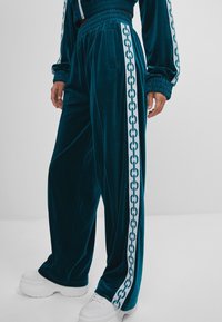 Tealové sametové tepláky mají boční kapsy a bílý vzor řetízkového spoje podél vnějších švů, doplněné robustními bílými teniskami.