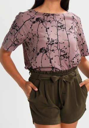 Mujer con un top malva con patrones de ramas negras y pantalones cortos de cintura paperbag verde oliva con cinturón atado, manos en los bolsillos.