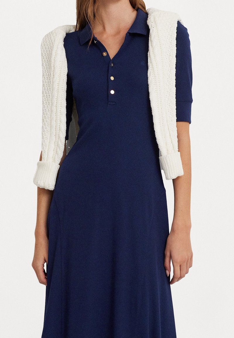 Námořnická modrá polo šaty s přiléhavým korzetem a volánovou sukní, s krátkými rukávy a zlatými knoflíky; krémový pletený svetr je přehozený přes ramena.