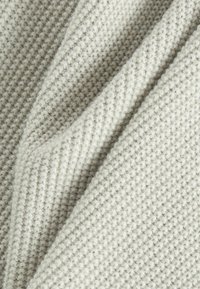 Tejido de punto suave color crema con un patrón texturizado similar a una gofrera y pliegues visibles que crean sombras.