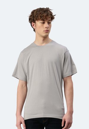 Junger Mann mit lockigem Haar, der ein schlichtes, hellgraues T-Shirt mit kurzen Ärmeln trägt und nach links blickt vor einem schlichten, hellen Hintergrund.